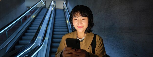 Happy smiling young woman standing on escalator going down holding smartphone in both hands chatting