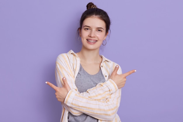 Happy smiling young woman wearing white shirt, having hair bun, pointing with index fingers to both sides, woman standing against lilac wall, looks at camera, expresses positive