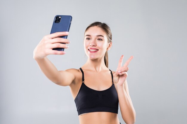 Счастливая sporty женщина делая selfie на изолированном smartphone на белой стене