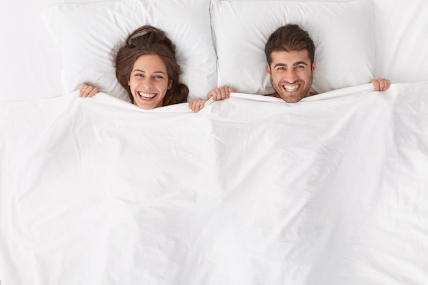 Free photo happy spouses enjoy spending time together, lie under white blanket, have positive expressions and smiles, stay in bed, awake after sleep or nap early in morning feel renewed after deep healthy night,