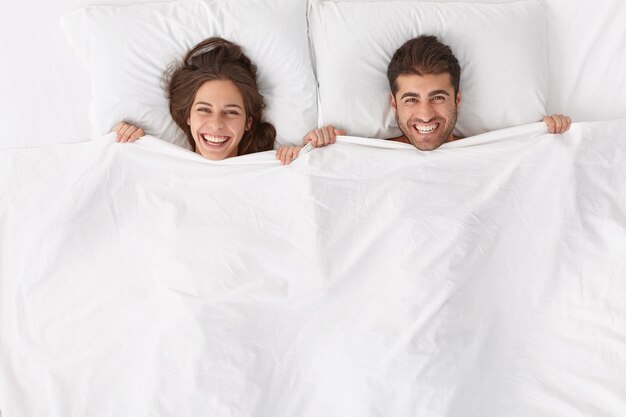 Happy spouses enjoy spending time together, lie under white blanket, have positive expressions and smiles, stay in bed, awake after sleep or nap early in morning feel renewed after deep healthy night,