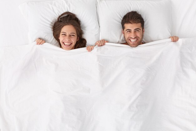 Счастливые супруги с удовольствием проводят время вместе, лежат под белым одеялом, имеют позитивные эмоции и улыбки, остаются в постели, бодрствуют после сна или дремлют рано утром, чувствуют себя обновленными после насыщенной здоровой ночи