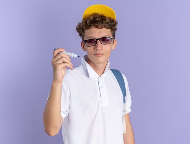 Free photo happy student guy in white polo shirt and yellow cap wearing glasses with backpack holding pen looking at it with smile on face