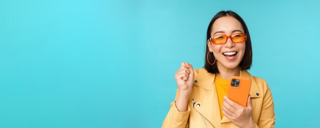 스마트폰을 사용하고 파란 배경 위에 서 있는 카메라를 보며 웃고 있는 행복한 세련된 아시아 소녀