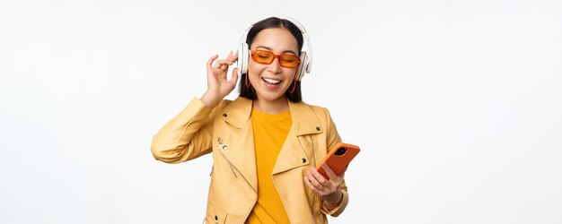 白い背景の上に立ってスマートフォンの歌と踊りを保持しているヘッドフォンで音楽を聴くサングラスで幸せなスタイリッシュなアジアの女性