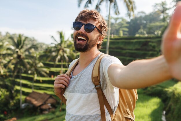 Happy stylish caucasian man with backpack travel in rice plantation and making self portrai for memories.