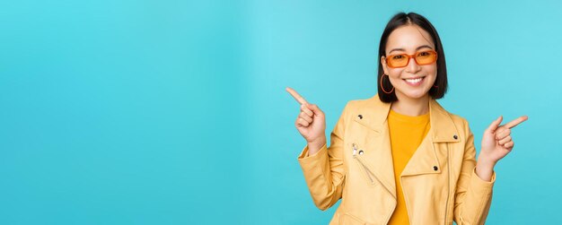 Happy stylish chinese girl in sunglasses points fingers sideways invites to check out see store discount shows left and right stands over blue background