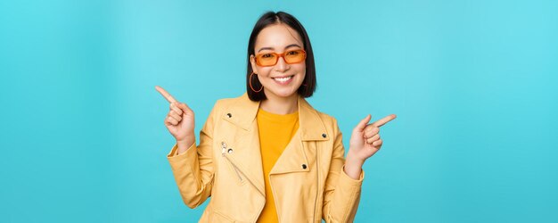 Happy stylish chinese girl in sunglasses points fingers sideways invites to check out see store discount shows left and right stands over blue background