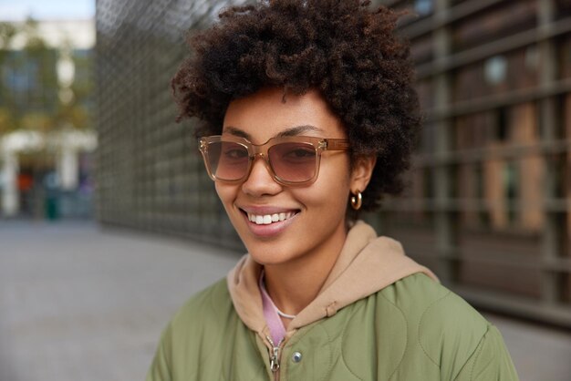 Happy stylish curly haired woman wears trendy sunglasses jacket and hoodie smiles gladfully walks outdoors poses against blurred background enjoys spare time People lifestyle and style concept