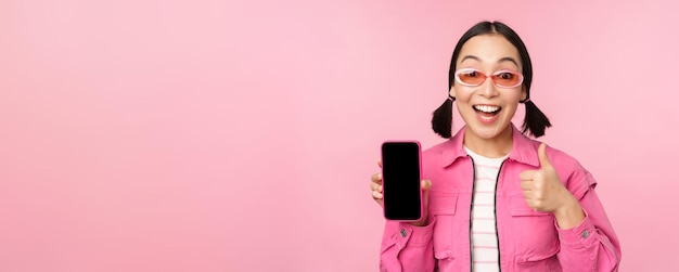 幸せなスタイリッシュな女の子は携帯電話でのアプリケーションをお勧めしますスマートフォンの画面とピンクの背景の上に立って親指を示す笑顔のアジアの女性モデル