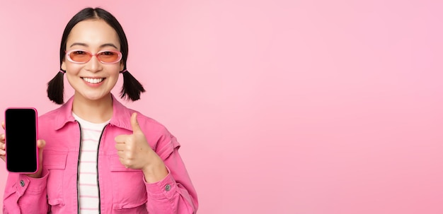 Happy stylish girl recommends application on mobile phone smiling asian female model showing smartphone screen and thumb up standing over pink background