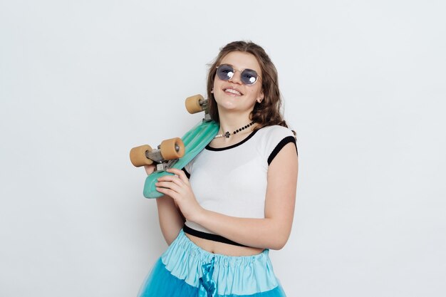 Счастливая стильная девушка-подросток в солнцезащитных очках позирует с пенни-доской на белом