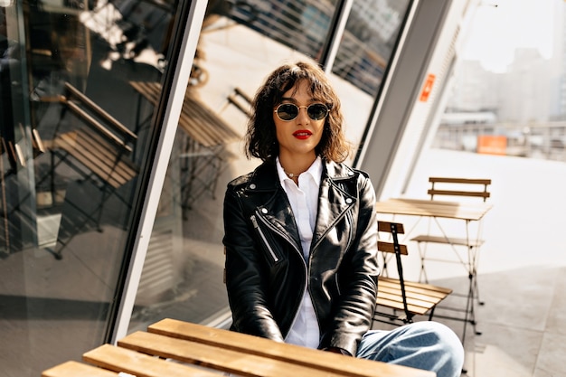 Happy stylish girl with short hair wearing glasses and leather jacket with red lips resting on summer terrace in sunlight