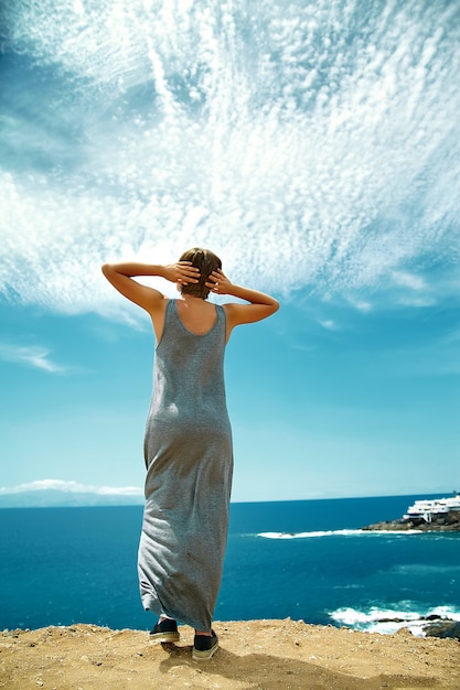 Happy stylish girl woman in casual hipster clothes standing on the cliff of mountain with her hands behind head on blue sky background