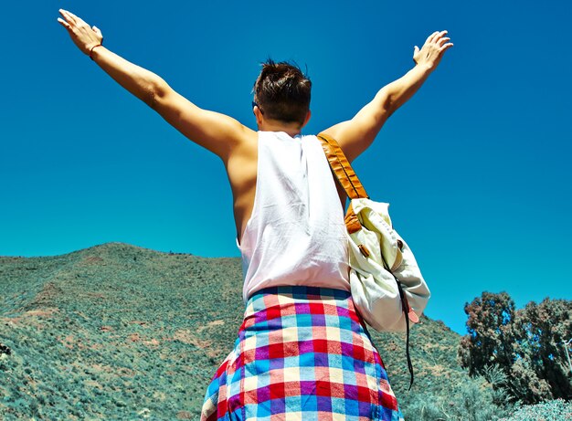 Happy stylish man in casual hipster clothes jumping in front of mountain with raised hands to the sun and celebrating success