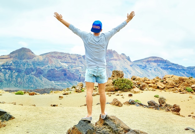 Happy stylish man in casual hipster clothes standing on the cliff of mountain with raised hands to the sun and celebrating success