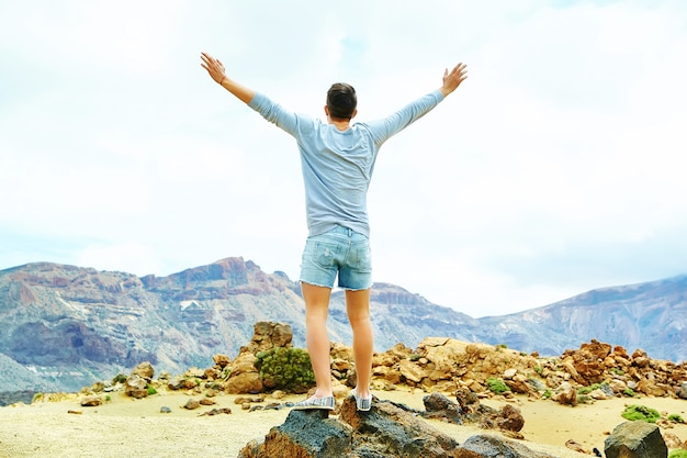 Happy stylish man in casual hipster clothes standing on the cliff of mountain with raised hands to the sun and celebrating success