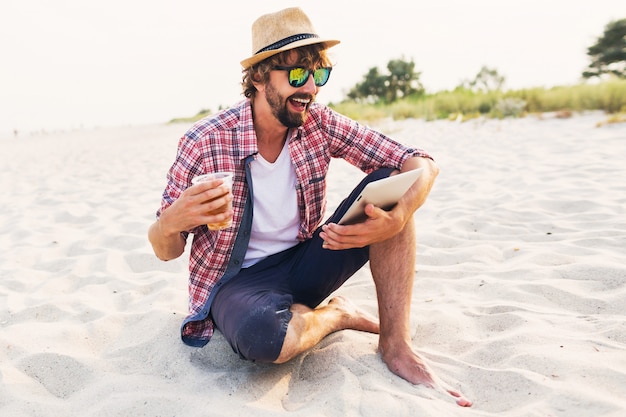 Happy stylish man using tablet and drinking beer on the beach