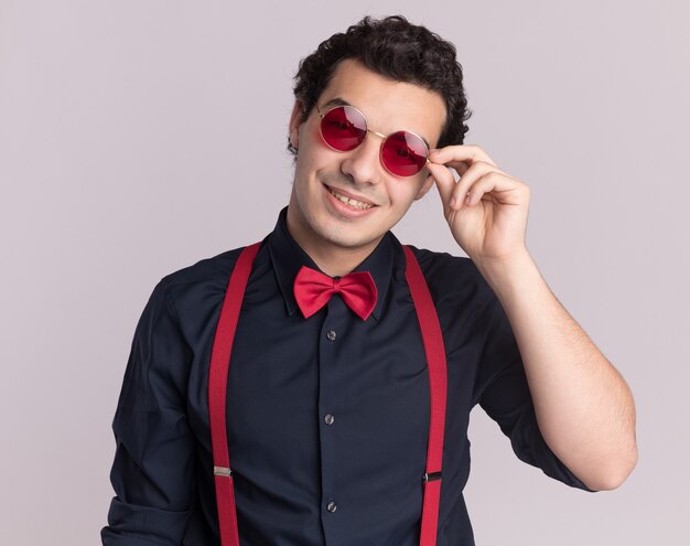 Happy stylish man with bow tie wearing glasses and suspenders looking at front smiling cheerfully standing over white wall