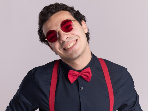 Happy stylish man with bow tie wearing glasses and suspenders looking at front with big smile on face standing over white wall