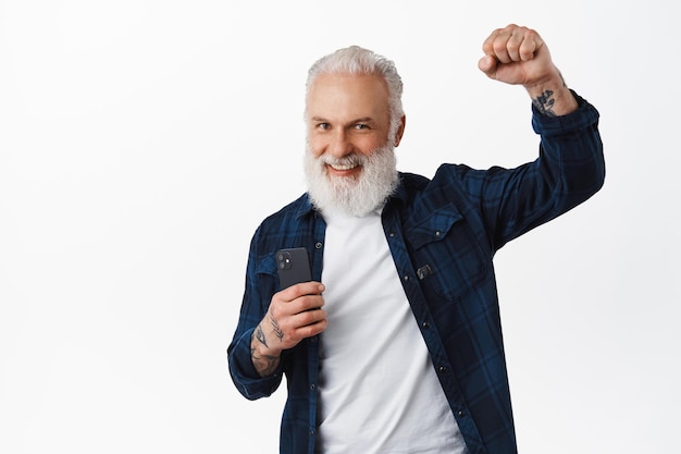 Happy stylish senior man holding smartphone and raising hand fist up in triumph, celebrating victory, winning money online, achieve goal, standing over white background