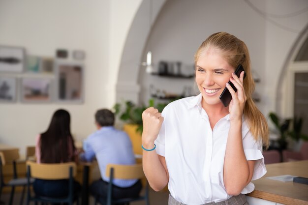 Happy successful beautiful young woman talking on mobile phone, making winner gesture, standing in co-working space