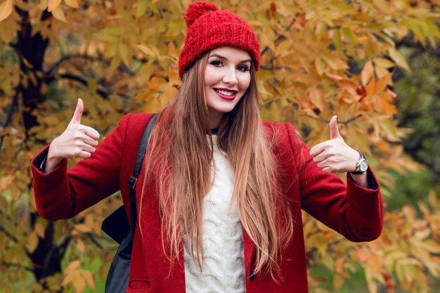 Happy successful blonde woman in red hat and jacket posing in autumn park.