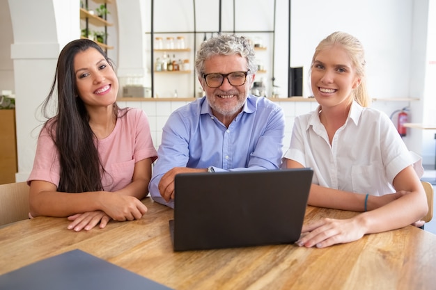 Happy successful business team sitting at table with open laptop, looking at camera, posing and smiling