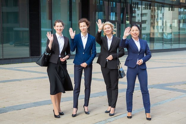 Счастливая успешная женская профессиональная команда, стоящая вместе возле офисного здания, размахивая приветом, позирует, глядя в камеру и улыбаясь. Полная длина, вид спереди. Концепция группового портрета деловых женщин