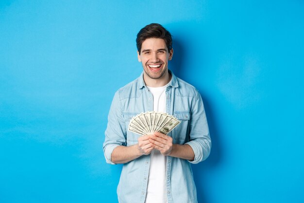 Happy successful man smiling pleased, holding money, standing over blue background