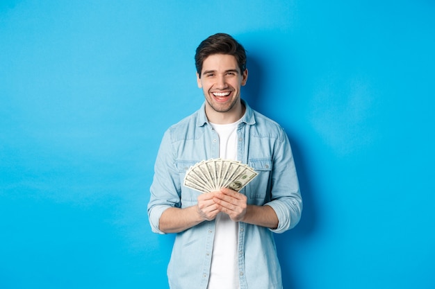 행복한 성공적인 남자는 만족스럽게 웃고, 돈을 들고, 파란색 배경 위에 서 있습니다.