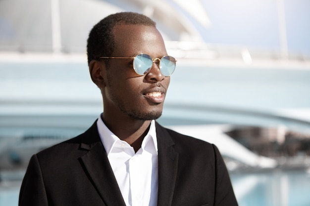 Happy successful young black employee in stylish formal wear and sunglasses looking cheerful, rejoicing at his career goals