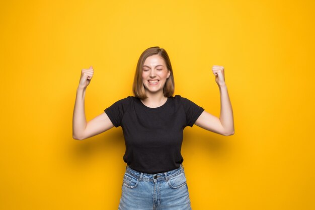 Happy successful young woman with raised hands shouting and celebrating success over yellow wall