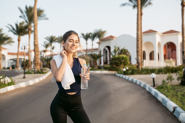 Happy sunny morning moments of joyful amazing young woman in sportswear smiling with bottle of water on street with palm trees. Training, workout, expressing positivity.