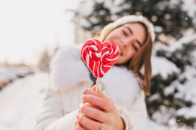 Happy sunny winter morning of joyful woman holding closeup pink heart lollypop on street. Sweet time, delicious, cold weather, snow, brightful emotions, having fun