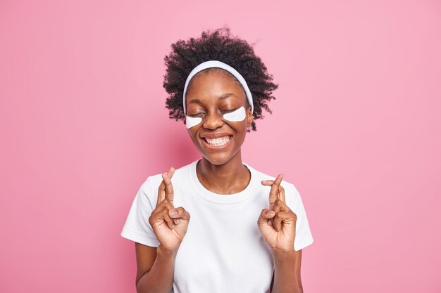 Happy superstitious millennial woman with dark skin curly hair crosses fingers makes wish keeps eyes closed applies patches for skin care 