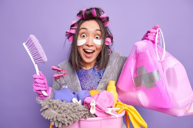 Happy surprised Asian woman applies hair rollers for making hairstyle patches under eyes olds trash bag cleaning brush poses near laundry basket dressed in robe isolated over purple wall