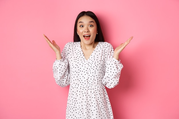 Happy and surprised asian woman rejoicing, spread hands and gasping amazed, looking with excitement and disbelief, standing over pink background