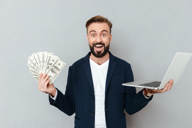 Счастливый удивленный бородатый человек в деловой одежде, держа деньги и портативный компьютер, глядя на камеру над серым