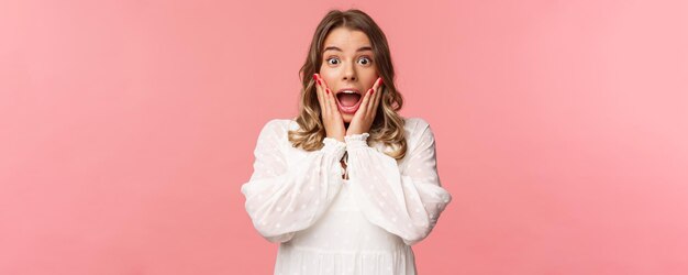 Happy and surprised blond girl hear awesome news gasping speechless staring camera with hands on cheeks being impressed and stunned over great event standing pink background