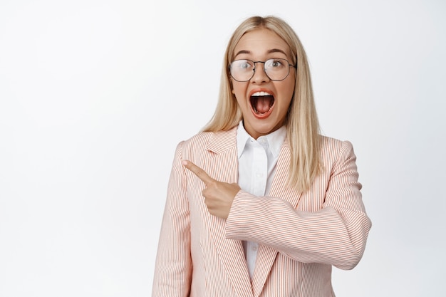 Free photo happy and surprised business woman pointing left, scream from excitement on white.