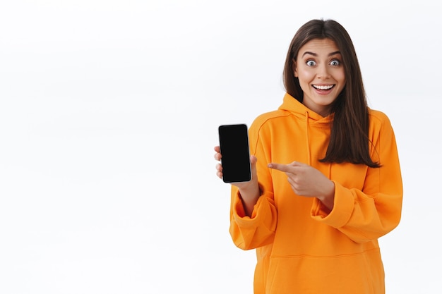 Happy surprised cute brunette girl in orange hoodie, holding mobile phone and pointing at display to show you awesome event going nearby, want book tickets, looking at you imrpessed