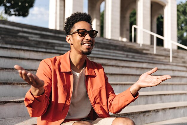 Happy surprised darkskinned man in orange jacket sunglasses and white tshirt shrugs smiles Charming guy sits on stairs outside