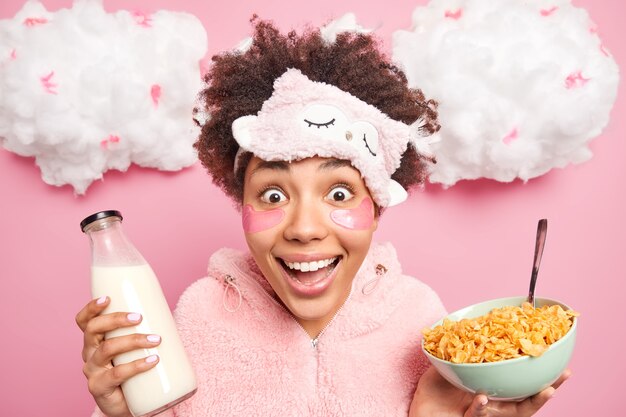 Happy surprised ethnic woman with Afro hair looks gladfully at camera holds bowl of cornflakes with milk wears nightwear stares positively at you enjoys healthy breakfast and beauty procedures