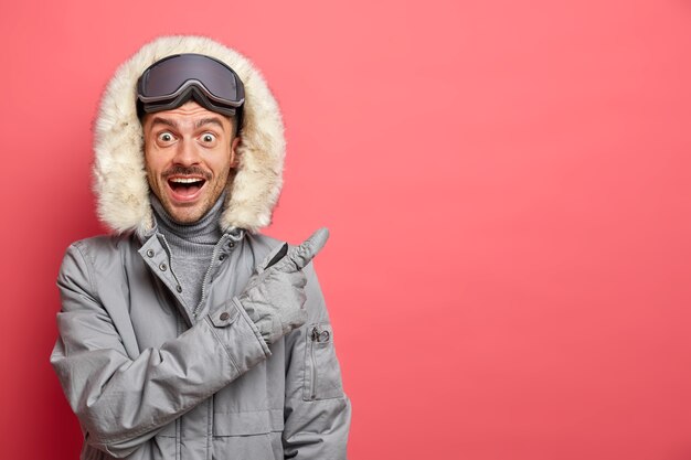 Happy surprised excited man snowboard rider in winter jacket relaxes after skiing has active day of recreation points away on blank space.