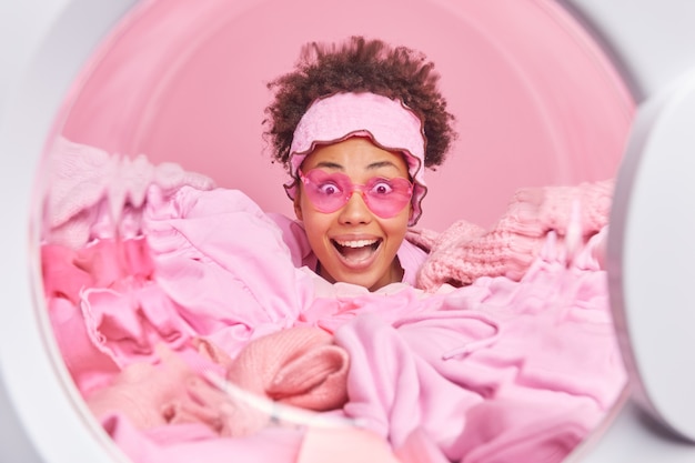 Foto gratuita la governante femminile sorpresa felice con hiar riccio indossa occhiali da sole a forma di cuore rosa infila la testa attraverso la pila di bucato