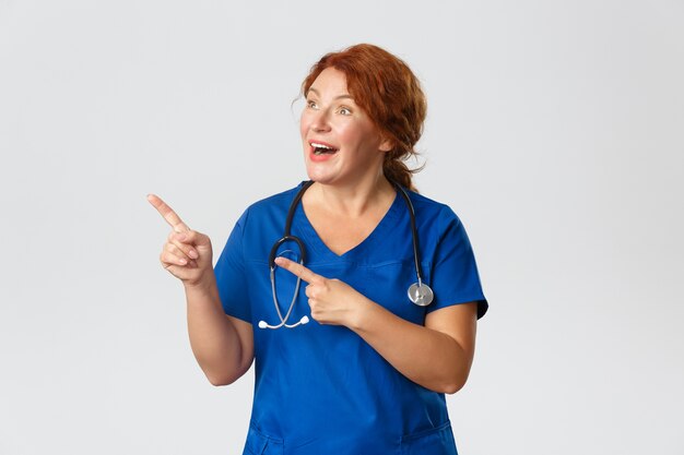 Happy surprised middle-aged doctor, female nurse in scrubs react to something wonderful or cute, pointing and looking upper left corner with smile