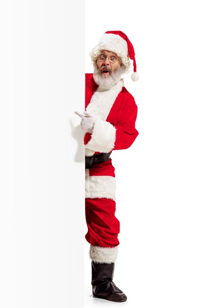 Счастливый удивлен Санта-Клаус указывая на пустой рекламный фон баннера с копией пространства. Улыбающийся старший мужчина показывает белый бланк пустого плаката