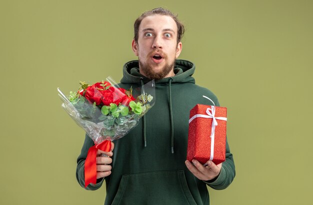 꽃 부케와 캐주얼 옷에 행복하고 놀란 젊은 남자와 녹색 배경 발렌타인 데이 개념 위에 서 그의 여자 친구를위한 선물
