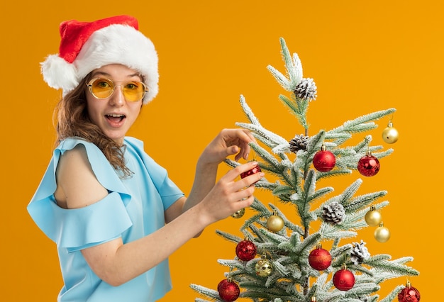 青いトップとオレンジ色の背景の上に立っているクリスマスツリーを飾る黄色いメガネをかけているサンタ帽子の幸せで驚きの若い女性